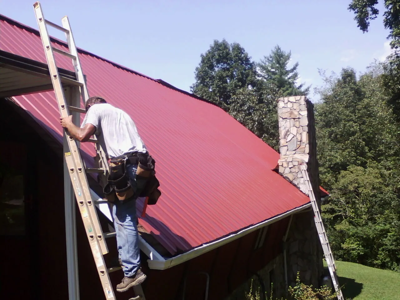 Roofer on a ladder beside a red metal roof and stone chimney — Soffit & Fascia Repair by Roofing Brothers in Shoshone